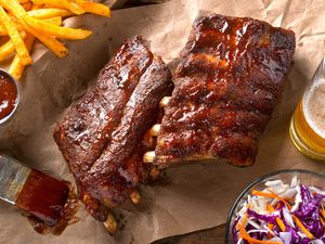 rack of glazed ribs on parchment paper at a table setting with a basket of fries, bowl of coleslaw, and a glass of beer 