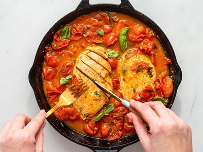 hands with knife and fork slicing cooked chicken breasts in a tomatoey sauce in a cast iron skillet