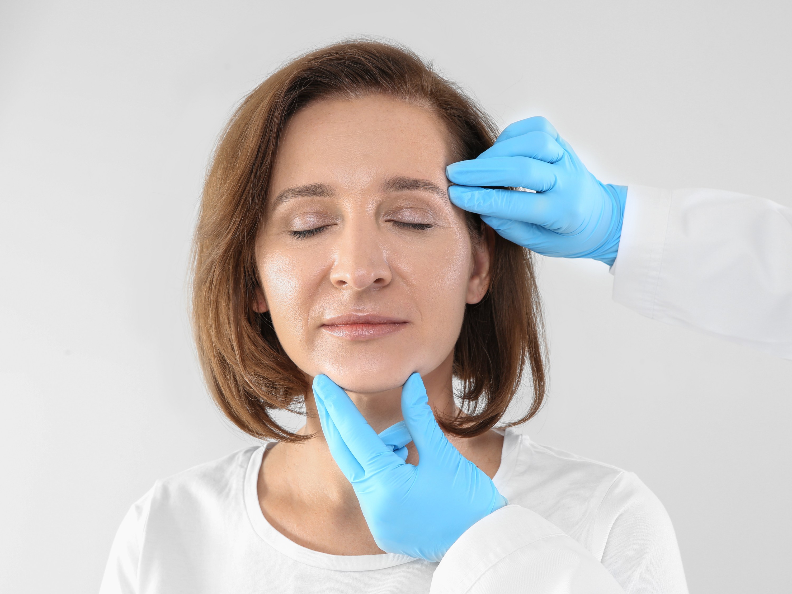 Dermatologist's gloved hands examine the face of a woman whose skin is getting more mature.