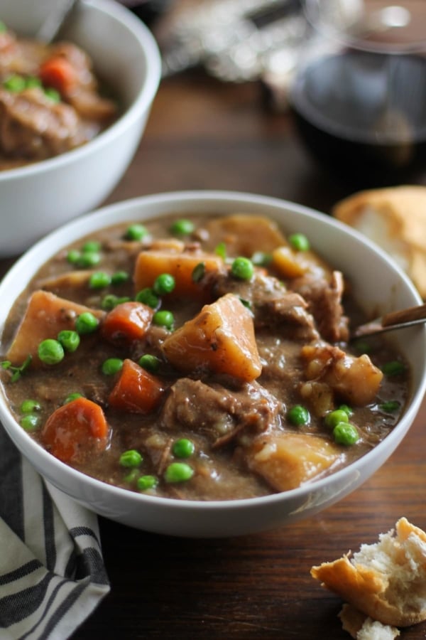 two white bowls of hearty beef stew on a rustic wood background with bread and wine to the side.