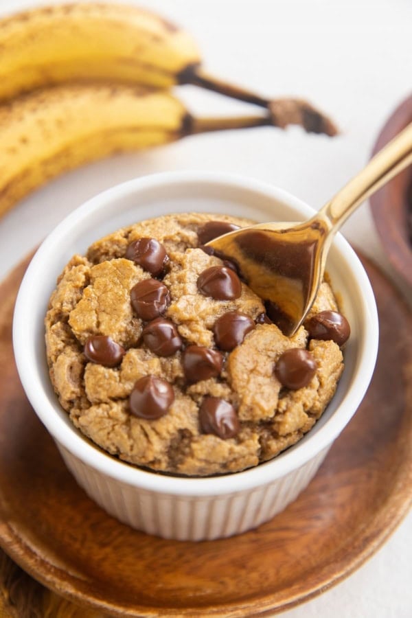 Ramekin of baked oats with chocolate chips on top and a golden spoon with ripe bananas in the background