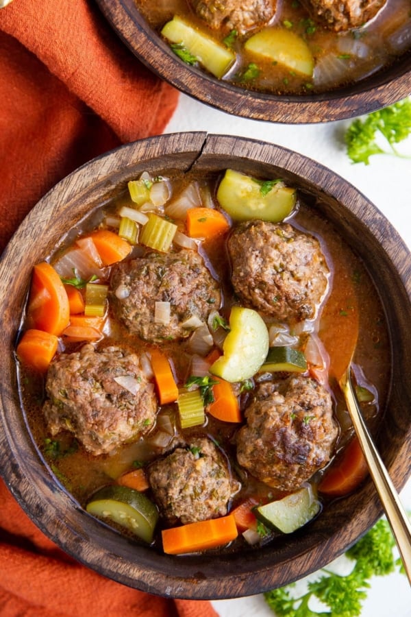 Two bowls of Albondigas soup in wooden bowls with a red napkin to the side and fresh parsley