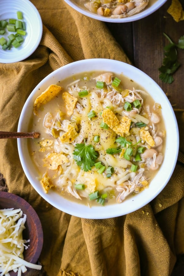 One large bowl of white chicken chili with chili toppings. A bowl of grated cheese to the side and a golden napkin.