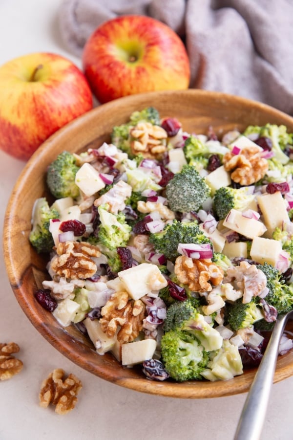 Wooden bowl of broccoli salad with a serving spoon, and fresh apples in the background.