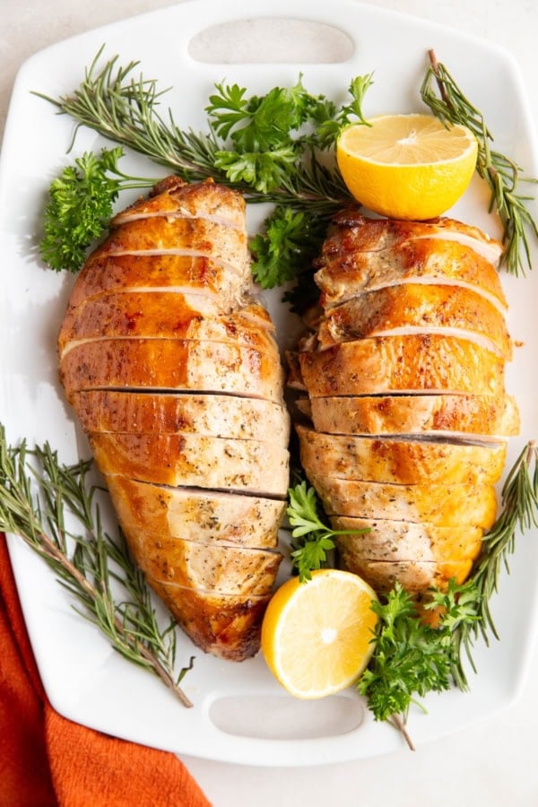 Garlic herb turkey breasts on a white serving platter surrounded by fresh herbs and lemon.