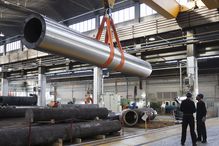 Steel factory workers in hardhats standing near a large steel tube hanging from an overhead crane.