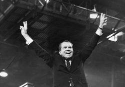 Black and white picture of Richard Nixon raising arms up and making "peace" signs with his hands