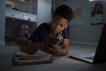 Teenage boy with headphones listening to music, using cell phone and doing homework on bedroom floor