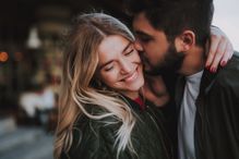 Precious moments of love. Close up portrait of handsome bearded guy kissing his girlfriend in cheek while she hugging him. Lady closing eyes with pleasure and smiling