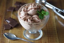 Chocolate ice cream in a dish with a sprig of mint on a wooden table with scattered chocolate bits.