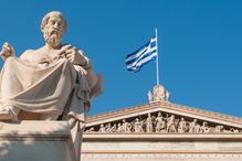 Statue of Plato in front of a building with the Greek flag against a blue sky.