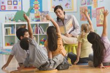 Teacher reading book in classroom, children (2-7) raising hands