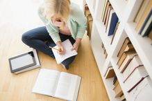 a woman sitting on the floor writing in a notebook with a book in front of her