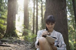 Woman writing in journal against tree in woods