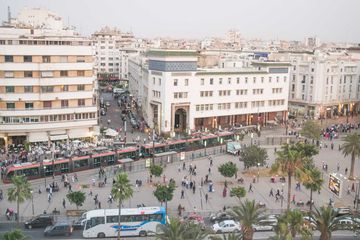 Avenue Moulay Hassan I with commercial buildings including a sidewalk cafe and shops. People walk in square. The network provides access between the city centre and the seaside suburb of Ain Diab and the Casa Voyageurs Station