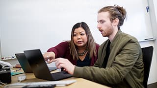 A professor and student both point to a laptop screen and discuss.