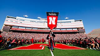 Paul Pechous carries the N U gonfolon into Memorial Stadium during commencement with bright blue sky in the background.