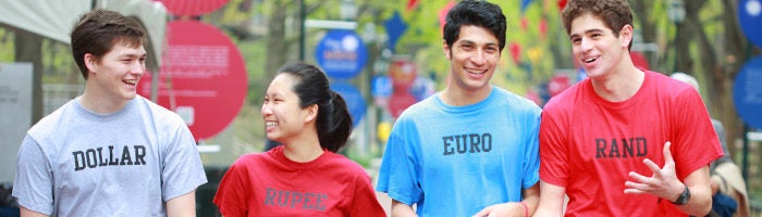 A small group of four people walking outdoors, each wearing a t-shirt with the name of a currency printed on it: Dollar, Rupee, Euro, and Rand. They appear to be engaged in conversation and smiling.