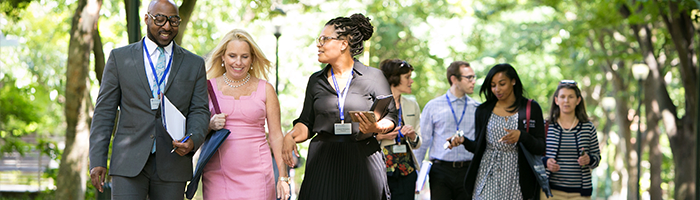 A group of people walking outdoors in a sunny, tree-lined park setting. They are engaged in conversation and carrying folders or electronic devices.